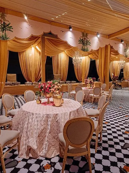 A dining area with elegant round tables set on a black-and-white checkered floor. The warm golden drapery and crystal chandeliers create a luxurious and inviting atmosphere for the wedding feast.