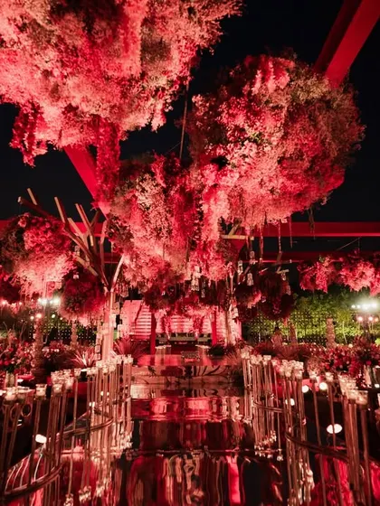 A stunning overview of the all-red mandap and dining setup. The mirrored aisle reflects the dramatic floral canopy and candlelight, doubling the impact and creating an immersive experience of royalty and romance for every guest.