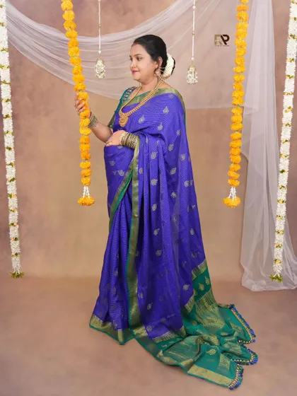 A full-length solo portrait in a vibrant blue and green saree. The marigold garlands add a festive touch to this traditional studio setup.