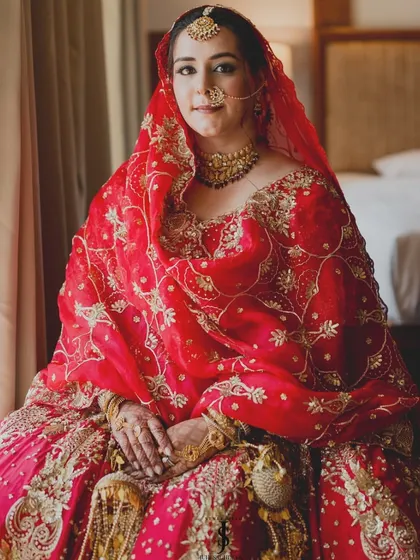 Seated by a window, the natural light beautifully highlights the rich color and gold embroidery of her traditional red bridal lehenga.