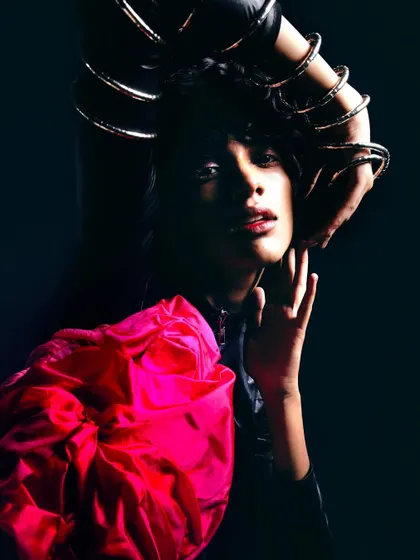A high-contrast studio shot where the model is framed by her own arms, adorned with bangles. The bright pink fabric pops against the deep black background, creating a visually arresting image.