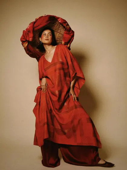 A dramatic, high-fashion pose showcasing a flowing red kaftan. The use of a traditional hat as a prop adds an artistic and sculptural element to the image.