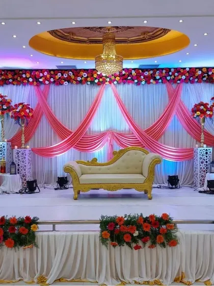 For this banquet hall reception, I created a vibrant and full stage setup. The backdrop features pink and white drapes with a thick floral border of red and orange blooms, complemented by tall floral arrangements on illuminated pedestals.
