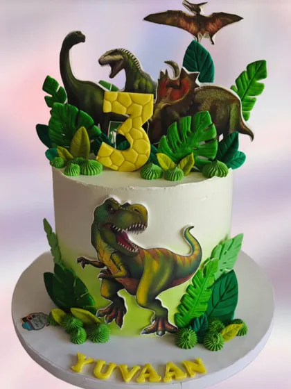 A tall dinosaur cake with realistic-looking toppers and lush green foliage. The design creates a dramatic and impressive centerpiece for a 3rd birthday party.