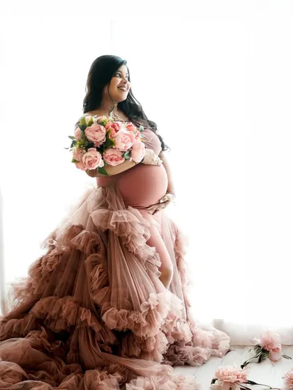 A high-key portrait of a glowing mother-to-be in a dramatic ruffled gown, holding a bouquet of pink peonies.