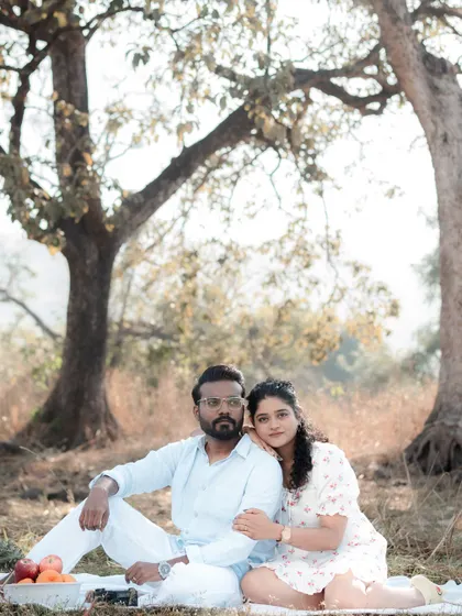 A serene, romantic picnic-themed pre-wedding shoot, with the couple sitting together under large trees in a sunlit field. This shows how we can create a beautiful, story-driven session in a natural outdoor setting.
