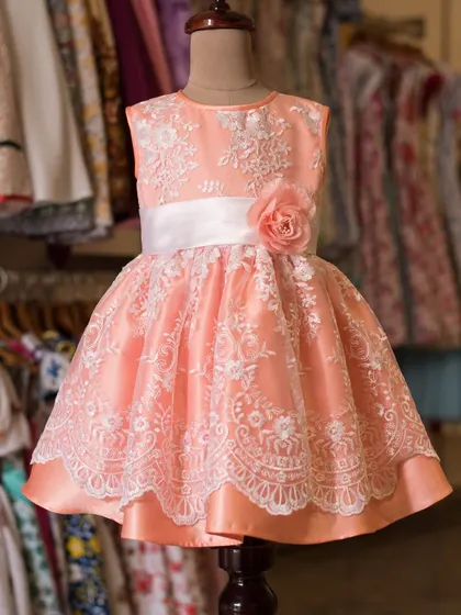 This peach-colored dress is adorned with exquisite white lace embroidery for a truly elegant feel. A white satin sash with a delicate rose completes this charming party look.