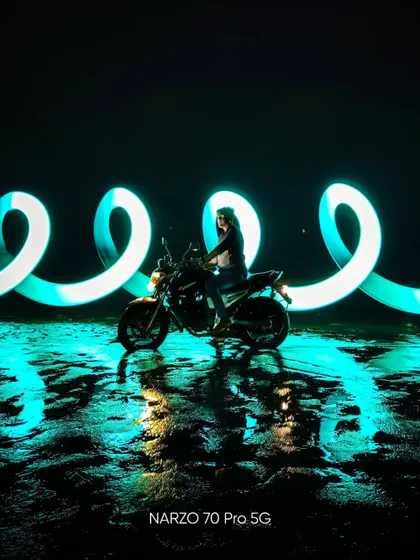 A creative portrait using light painting behind a motorcycle at night. The swirling blue light creates a futuristic, energetic mood, captured with the Narzo 70 Pro 5G.