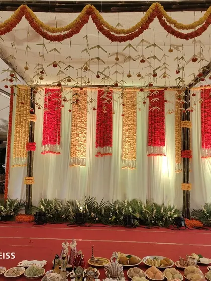 A traditional stage backdrop for a ceremony, using hanging garlands of red and yellow marigolds. The ceiling is decorated with woven palm leaves and marigold strings, creating a festive, ethnic look.