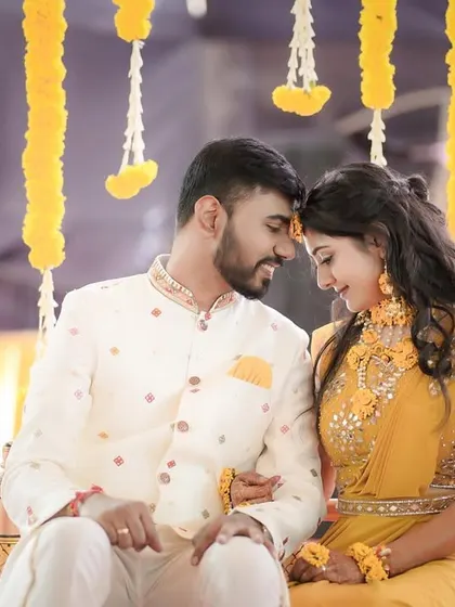 A classic Haldi ceremony portrait. The couple sits together, foreheads touching, under a canopy of hanging marigolds, perfectly capturing the essence of the ritual.