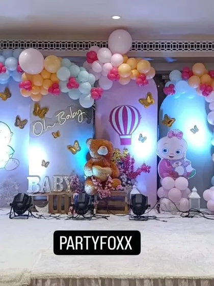 A cute baby shower theme with pastel balloon arches, character cutouts, a central teddy bear prop, and a hot air balloon detail.