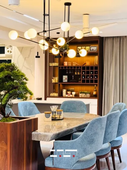 The dining area is designed for memorable meals, featuring a stunning marble-top table with a live bonsai centerpiece. The space is illuminated by a modern globe chandelier, with a custom bar cabinet in the background.