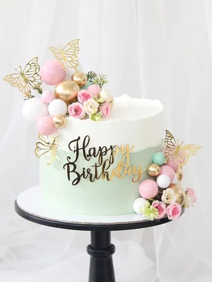 A sweet and simple birthday cake with a mint green and white ombre finish. The design is accented with clusters of pink and gold spheres, butterflies, and small flowers for a touch of whimsy.