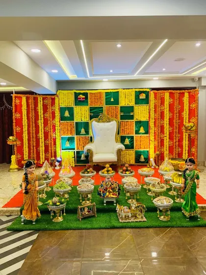 A wide shot of a traditional baby shower decoration in a spacious hall. The green, yellow, and red theme is carried through from the backdrop to the floor, creating a cohesive and festive look.