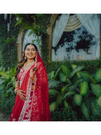 A happy bride in her red Parsi Ghara lehenga, standing in a beautiful garden setting. Her smile lights up the entire frame.