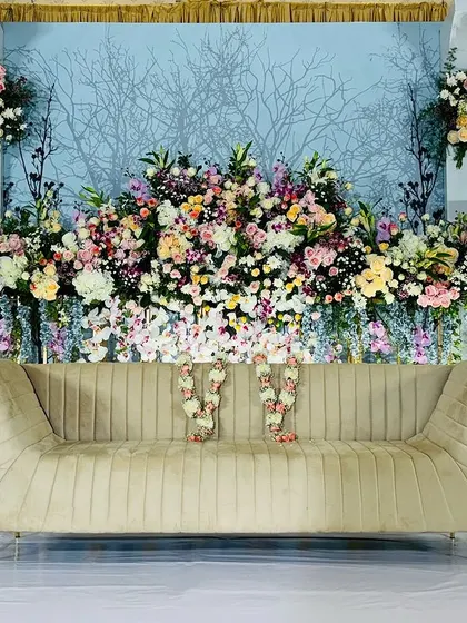 A full view of a majestic reception stage, where a pastel blue floral wall is flanked by multi-level white panels and golden accents. The design is elevated by beautiful chandeliers and lush foreground greenery.