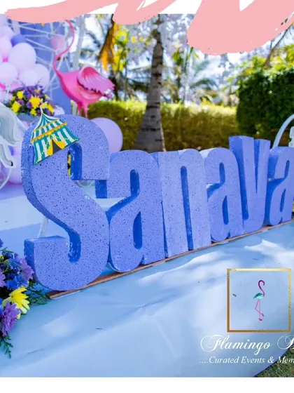 A large, glittering 'SANAYA' sign for the other birthday twin, surrounded by carousel horses and flowers.