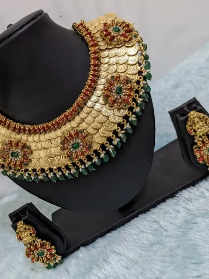 A unique neckpiece featuring a base of kaasu (coins) adorned with Jadau-style red coral and green stones. The matching earrings feature Goddess Lakshmi.
