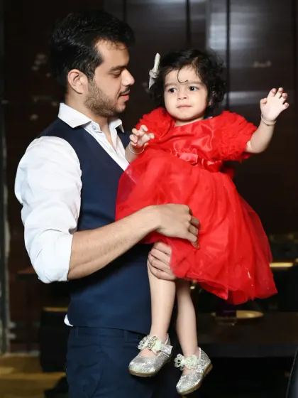 A father holds his daughter, dressed in a bright red party dress. We focus on capturing the connection between parents and their children during the celebration.