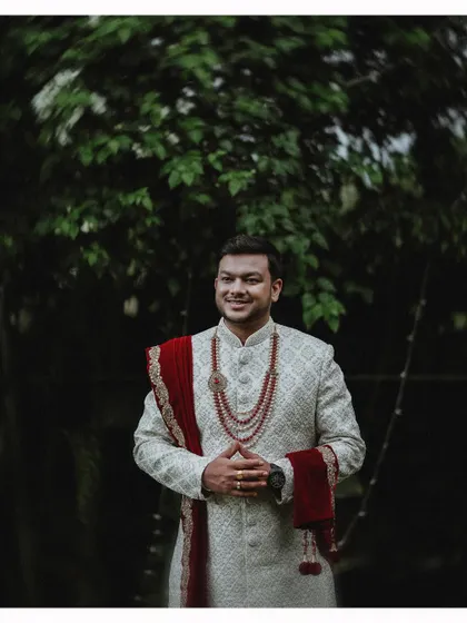 A handsome portrait of the groom in a white sherwani with a red stole, looking ready for his wedding.