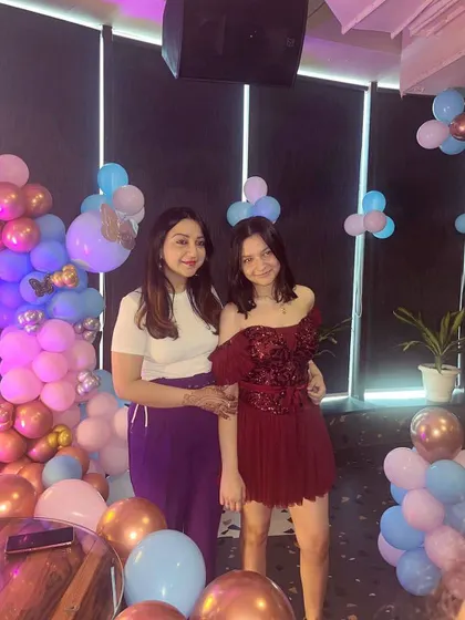 A happy client posing with her mother at her party. She looks radiant in her custom maroon off-shoulder dress, which features an applique bodice and a tiered, frilly skirt.