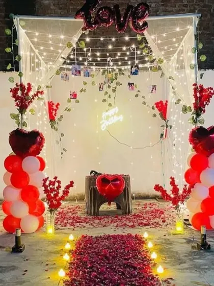 I design magical proposal setups like this one for lawns or terraces. This arrangement features a romantic canopy with fairy lights, a 'Happy Birthday' neon sign that can be customized, balloon columns, and a walkway of artificial rose petals and candles.