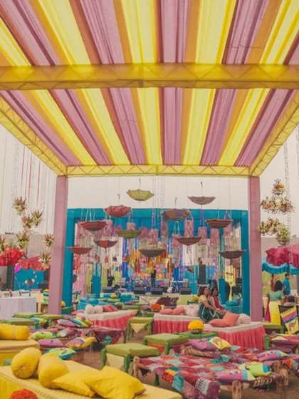 A vibrant and colorful Mehendi or Sangeet setup. The space is filled with low seating, colorful cushions, and hanging umbrellas, creating a festive and joyful atmosphere.