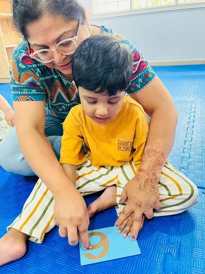 A teacher helps a child trace a sandpaper number, providing gentle guidance and encouragement in this early math activity.