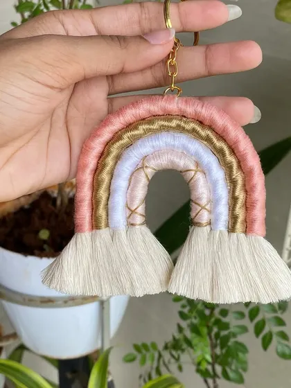A close-up of our handmade macrame rainbow keychain in a soft pink and gold colorway. The gold thread adds a little bit of sparkle, making it a stylish accessory for bags or keys.
