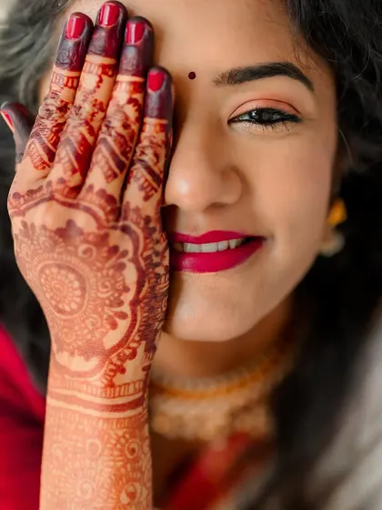 A close-up portrait of a bride, her face partially covered by her henna-decorated hand. Her smile and bright lipstick add a pop of color.
