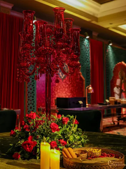 A striking red glass candelabra serves as a centerpiece on a guest table. The table is also adorned with red roses, candles, and a tray of traditional spices, continuing the theme of symbolic beauty.