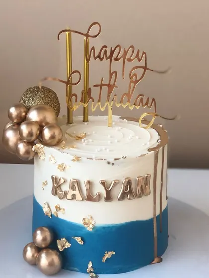 A stylish blue and white cake with a gold drip, balloon clusters, and a custom name.