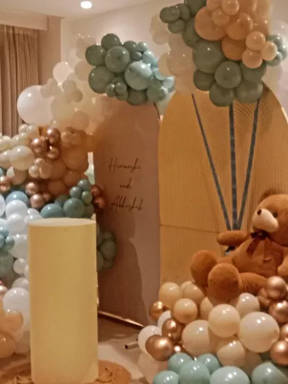 A closer look at a home-based baby shower setup. This design features multiple balloon arrangements in pastel blue, green, and beige, with a large teddy bear as the main focus.