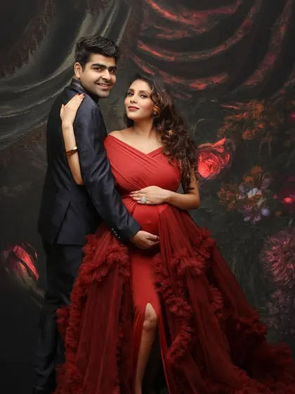 A sophisticated couple's portrait against a dark, floral-textured backdrop. The mom-to-be's vibrant red gown stands out beautifully, creating a photo that is both romantic and bold.