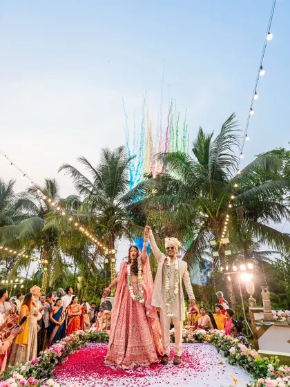 The couple raises their hands in celebration after the varmala, with colorful smoke bombs going off in the background. A vibrant and joyous moment.