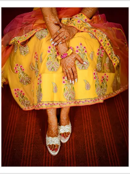 A top-down detail shot focusing on the bride's embroidered yellow lehenga, her henna-adorned hands, and her embellished footwear. I believe in capturing the beauty of the entire bridal ensemble.