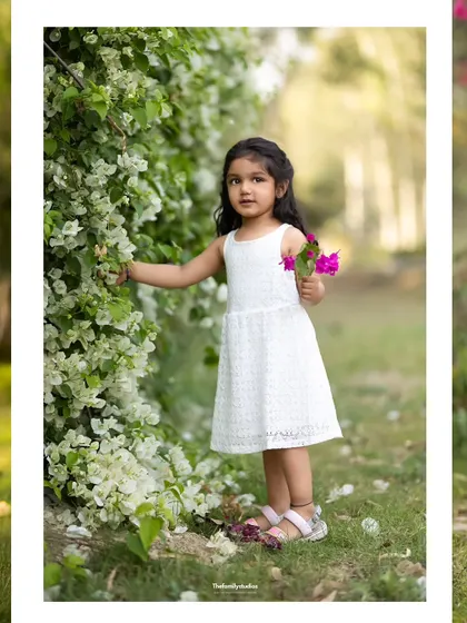 A young girl in a white dress stands by a wall of white flowers, creating a beautiful and serene outdoor portrait.