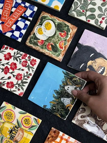 A hand holding up a tile with two little ghosts hiding in the leaves. We can't wait to see all 166 of these tiles installed on our studio wall.