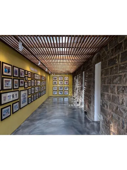 A gallery corridor where a rough, exposed stone wall of the original structure is juxtaposed with a smooth, yellow exhibition wall. Light filtering through the trellised ceiling creates rhythmic patterns, adding another layer of texture to the space.