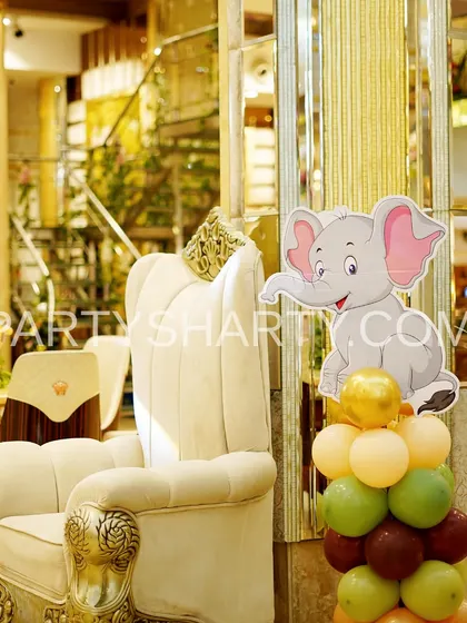 A cute elephant character prop on a balloon column, placed next to a regal throne chair for the birthday child in a jungle-themed party.