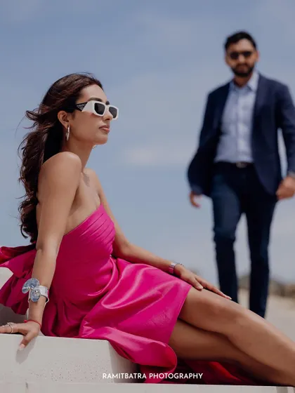 A close-up, fashion-forward shot of the bride in a pink dress and white sunglasses, with the groom blurred in the background.