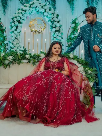 A stylish reception portrait. The groom stands beside his seated bride, whose elegant red gown flows around her, creating a grand and romantic image against the beautifully decorated stage.