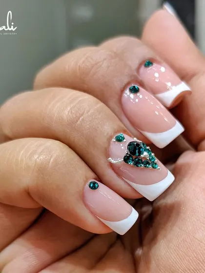 A full hand view of the French manicure with green crystal accents. The design is balanced, with a single crystal on some nails and a full cluster on the accent nail.
