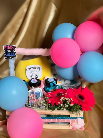 A vibrant gender reveal hamper decorated with pink and blue balloons. Inside the crate, I've included fresh flowers, a "Coming Soon" plush toy, and small gift jars to celebrate the parents-to-be.