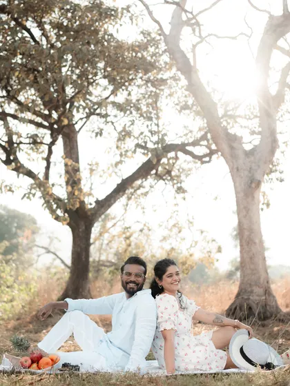 A happy couple sits back-to-back on a picnic blanket under trees, smiling brightly. The warm sunlight filtering through the leaves creates a beautiful, relaxed atmosphere for this outdoor shoot.