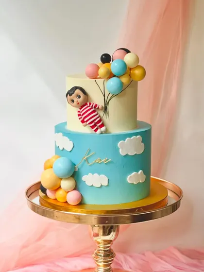 A whimsical two-tiered cake with a Cocomelon-style figure floating up with balloons. The ombre blue bottom tier is decorated with fluffy fondant clouds.