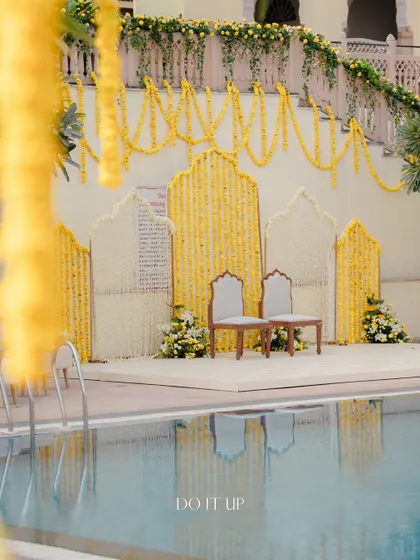 The poolside Haldi setup, with a backdrop of white and yellow floral panels. The reflection in the water adds another layer of beauty to the scene.