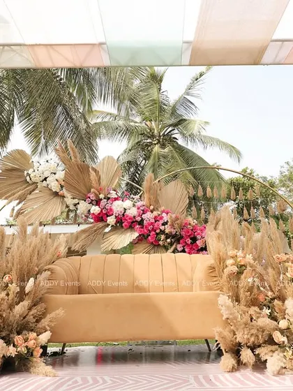 For a sun-drenched Mehndi ceremony, we designed this stunning boho-chic seating area. The arrangement features a comfortable lounge sofa framed by an arc of dried palm leaves, pampas grass, and vibrant pink florals, creating a picture-perfect spot for the couple.