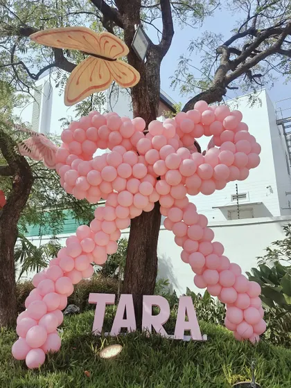 A giant pink balloon bow tied around a tree, personalized with the name "Tara." This is a creative and eye-catching way to add a personalized touch to an outdoor party space.