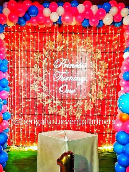 A 'Princess Turning One' birthday setup featuring a vibrant backdrop of fairy lights and a custom vinyl sign. The stage is framed by a colorful balloon arch in pink and blue, creating a festive space for a first birthday party.